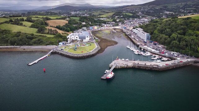 Aerial footage of coastal town and port of Bantry in West Cork Ireland with inner harbor marina boats moored along the pier and green rolling hills in the background.