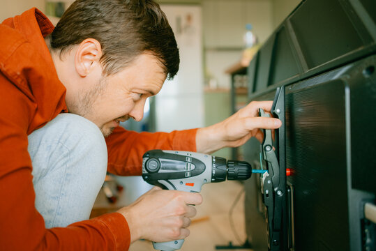 Man installing a large flat screen television onto a wall mount, carefully attaching the metal brackets to the back of the tv using a power drill, working on a home improvement project