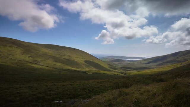 Time-lapse video of clouds passing over green mountain landscape and glacial valley of Conor Pass on Dingle Peninsula in Ireland, creating shifting shadows on rolling hills with distant bay.
