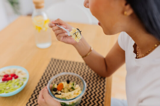 Young woman enjoying a healthy pasta salad breakfast at home. Relaxed young woman eating a nutritious homemade meal in a comfortable kitchen. Healthy eating and wellbeing concept.