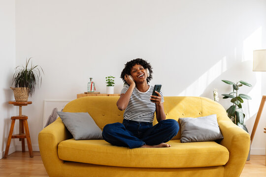 Happy African American woman enjoying online music with headphones while sitting on sofa at home
