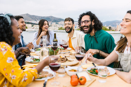 Diverse friends laughing and sharing a casual summer rooftop dinner, enjoying food, wine and city views while relaxing and celebrating togetherness on a terrace