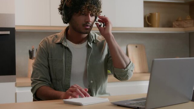 Concentrated Indian Arabian man student studying online at home kitchen wearing headset male watching webinar writing notes remote lesson education internet course educational video learning session