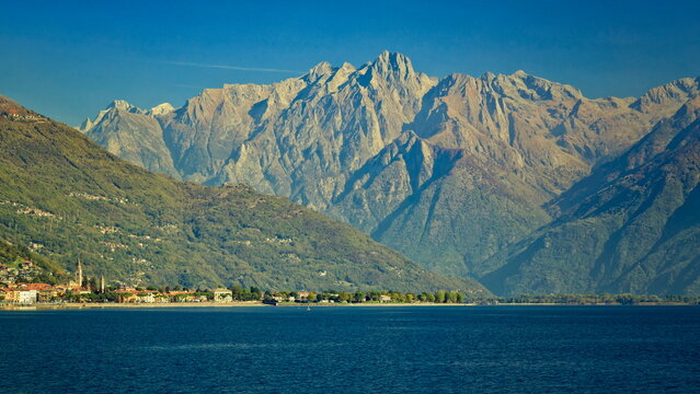 Gravedona ed Uniti, Italy - 17.10.2022: Jagged rocky Alps peaks towering over the deep blue Lake Como and a quiet lakeside town shore