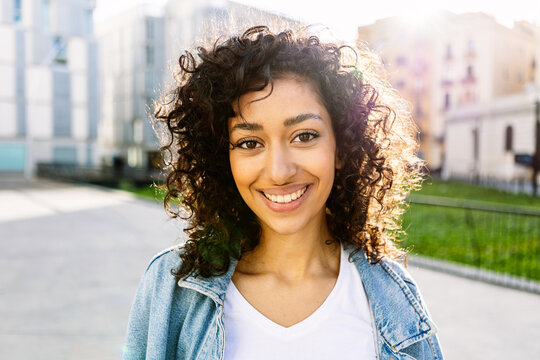 Portrait of young happy pretty woman with curly hair smiling at camera standing outdoors over urban background. Happiness and people concept