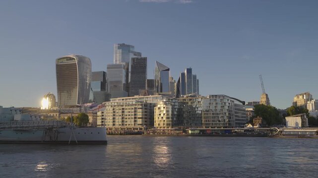 Low-angle view across the River Thames toward the City of London financial district skyline and historic HMS Belfast during early evening golden hour.