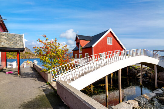 Picturesque white wooden bridge in the fishing village of Ure in Lofoten, surrounded by traditional red wooden houses, water and Nordic coastal scenery beneath a blue sky.
