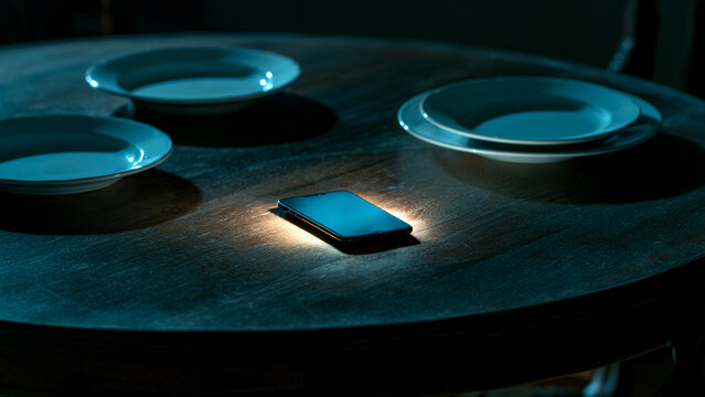 Smartphone with glowing blank screen lying on dark wooden dining table between empty plates in moody lighting. Concept addiction during meals, food delivery app, or digital diet