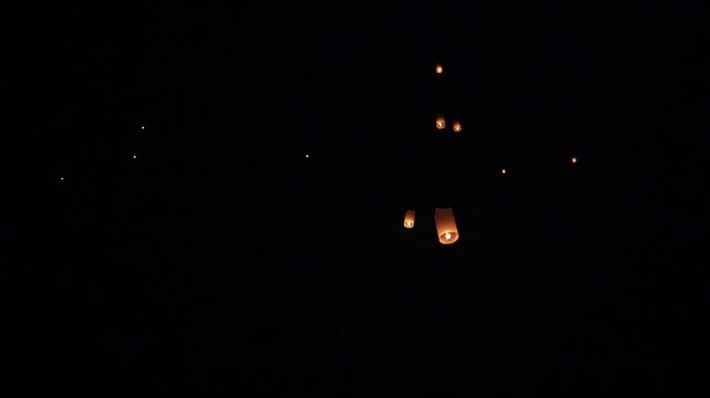 Warm glowing paper sky lantern rising at night with visible flame inside glowing thin paper and blurred lanterns in outdoor evening sky
