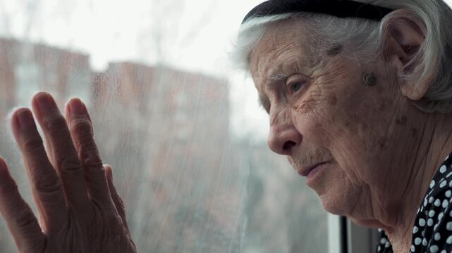 Pensive and lonely elderly woman 80 90 years with wrinkled hands and sad expression looking through window, experiencing melancholy while touching glass as it rains outside the nursing home