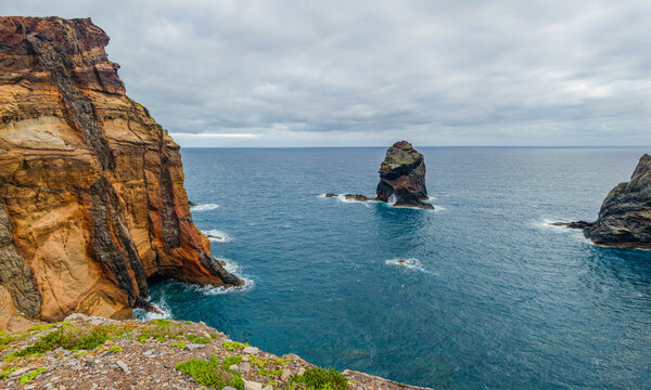 Tall golden-colored volcanic rock cliffs with the "gorilla head" rock framing a single rocky islet in the blue Atlantic ocean at Ponta de Sao Lourenco, Madeira, Portugal.