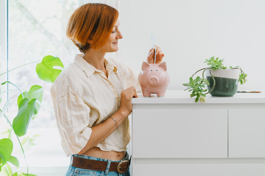 A mature caucasian woman carefully drops cash into a pink piggy bank while standing in her new home. Personal savings growth and mortgage payment preparation concept.