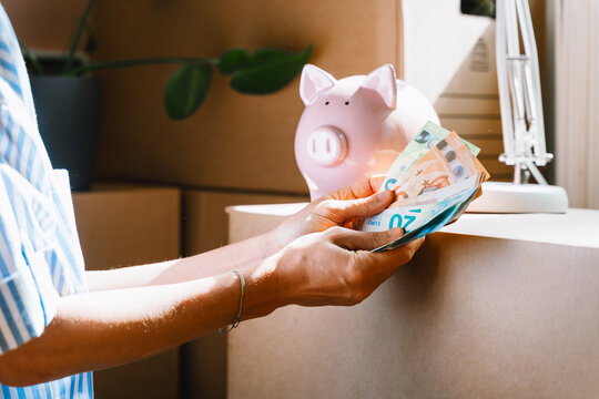 A person's hands carefully hold euro banknotes next to a pink piggy bank inside an apartment. Personal finance management and domestic home savings concept.