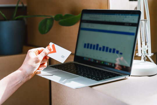 A person holds a credit card while analyzing financial charts on a laptop screen near moving boxes. Online banking and digital mortgage payment concept.
