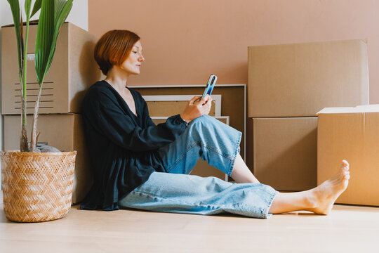 A mature caucasian woman sits on the floor among cardboard boxes using her smartphone device. Easy relocation process and modern digital lifestyle concept.