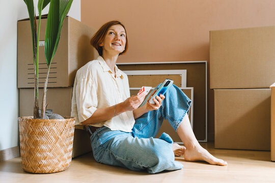 A mature caucasian woman holds a credit card and smartphone while sitting by unpacked moving boxes. Online shopping and remote household purchasing concept.