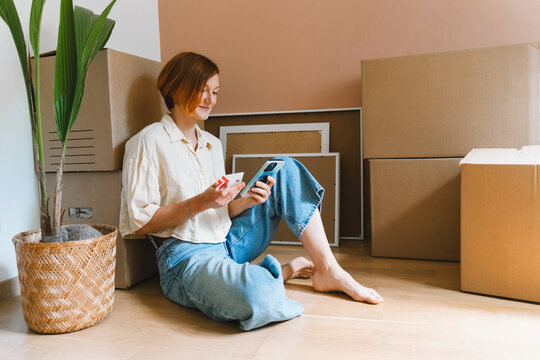 A mature caucasian woman holds a credit card and smartphone while sitting by unpacked moving boxes. Online shopping and remote household purchasing concept.
