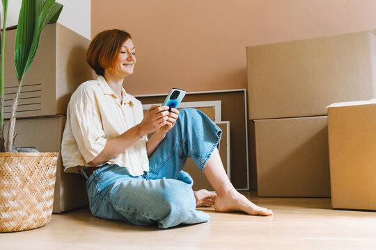 A mature caucasian woman sits on the floor among cardboard boxes using her smartphone device. Easy relocation process and modern digital lifestyle concept.