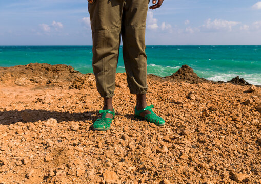 A person wearing green jelly sandals stands on a rocky coastline, Obock Region, Khor angar, Djibouti