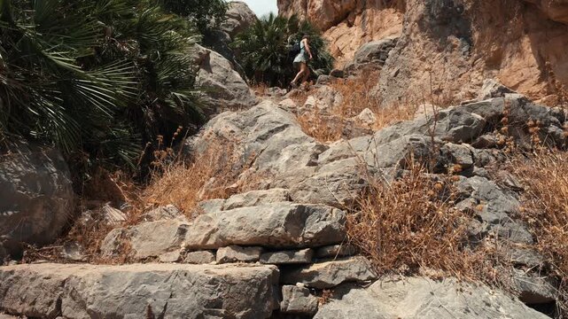 A solo female traveler climbing a rocky path flanked by palm fronds and dry brush in El Chorro Andalusia Spain under bright natural light on a rugged sunlit mountain trail