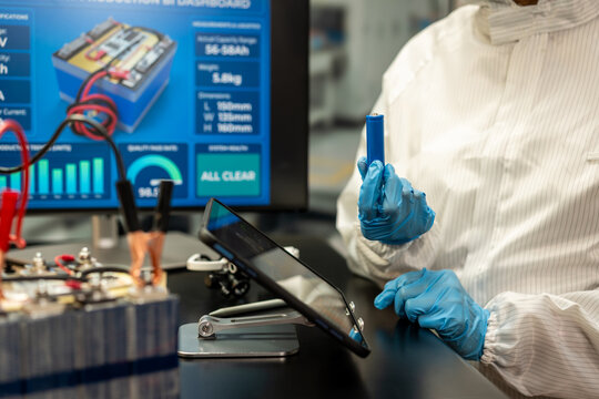 Technician wearing blue gloves holds a battery cell beside a tablet and electronic testing equipment in a modern laboratory.