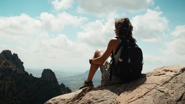 A young woman hiker sitting on a rocky ledge overlooking a mountainous valley in El Chorro Andalusia Spain under a partly cloudy sky with distant hills and trees
