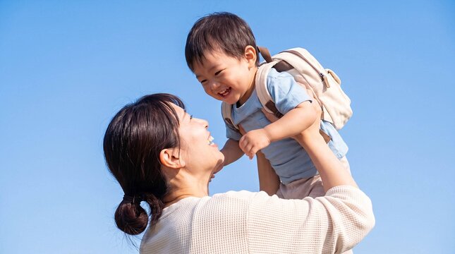 Mother lifting laughing toddler with backpack against blue sky, happy Asian family outdoors