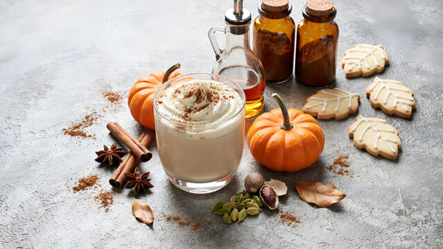 Pumpkin spice latte with whipped cream, cinnamon sticks, star anise, spices, and leafshaped cookies on a rustic gray background