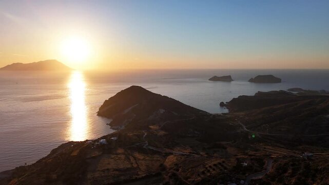 The low sun casts a bright reflection across the sea off Plaka on Milos, Greece, with dark headlands, offshore islets and terraced dry slopes filling the aerial panorama.
