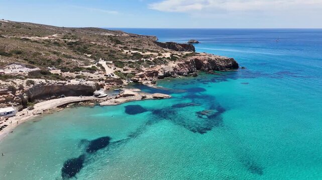 Clear turquoise water spreads across Paralia Firiplaka on Milos, Greece, as the camera moves above a sandy beach, scattered swimmers, dark seabed patches and rocky coast.