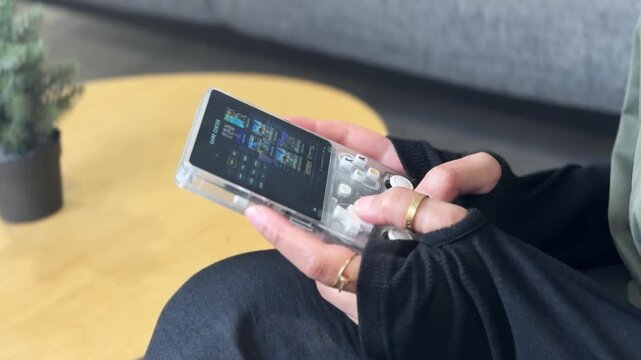 Close-up of a person's hands wearing gold rings holding a transparent handheld gaming console. The individual is relaxing indoors on a sofa near a wooden table with a small plant during daytime.