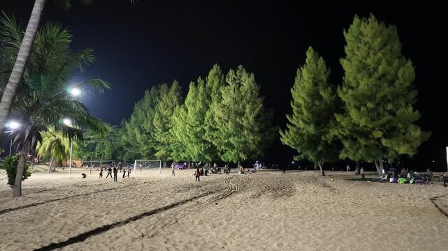 Slow motion wide shot of families enjoying a night picnic and children playing on the illuminated sand under pine trees at Pantai Puteri in Melaka, Malaysia.
