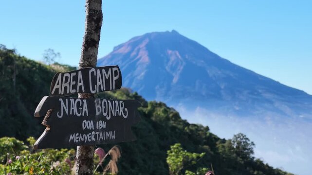 Naga Bonar campsite sign on Mount Kembang hiking trail with Mount Sumbing in the background, showcasing scenic mountain landscape and outdoor adventure in Central Java, Indonesia.