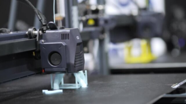 3D printer extrudes light blue plastic. Printer head moves slowly. Black platform holds forming object. Background shows blurred workshop equipment.