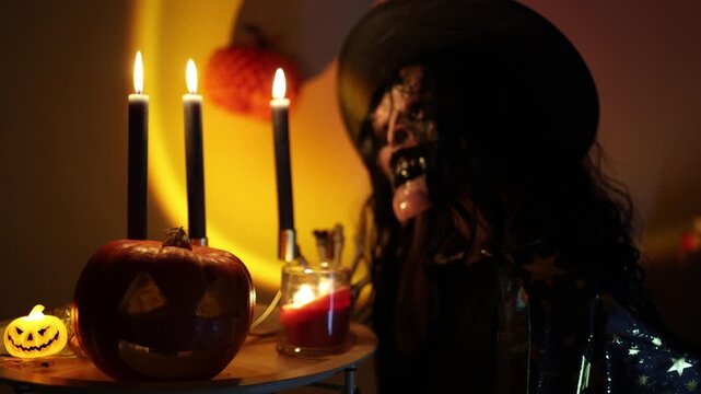 Halloween woman in skeleton makeup and witch costume poses beside carved pumpkin and glowing candles creating an eerie October celebration in warm dramatic lights indoors