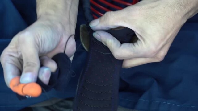 Artisan hand sews the padded edge of a kendo men mask using a bright orange fingertip thimble