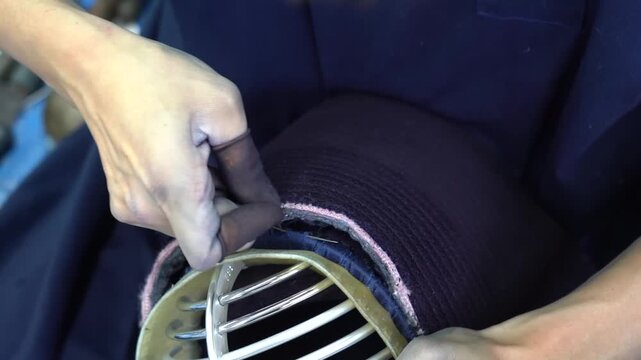 Artisan pushes a needle through thick indigo cotton along the grille rim of a kendo mask using a thimble