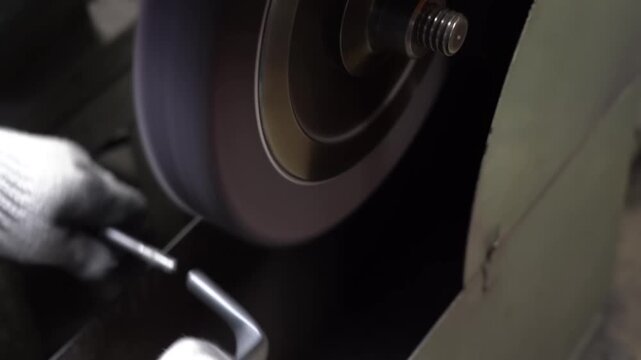 Gloved craftsman grinds a slender steel rod against a spinning abrasive wheel at a bench grinder