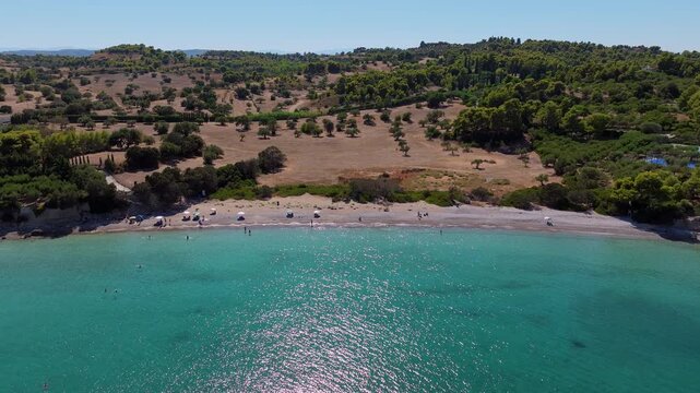 Cinematic drone shot of Paralia Kounoupiou Beach in Kranidi, Greece, showcasing crystal-clear turquoise waters, a secluded cove, and lush cypress-covered hillsides.