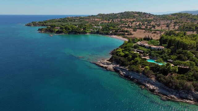 Aerial view of sunbathers enjoying the calm turquoise waters of Paralia Kounoupiou Beach in Kranidi, Greece, framed by olive groves and rolling coastal countryside.