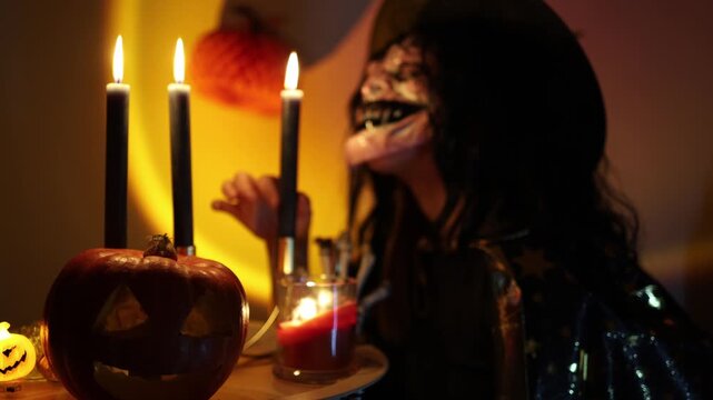 Halloween woman in skeleton face makeup raises her hand near glowing candles and carved pumpkin while posing in witch costume during a moody October celebration at home at night