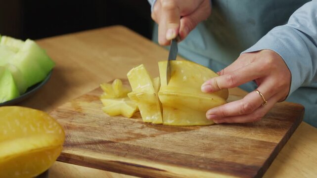 person cutting a fresh ripe yellow starfruit, also known as carambola, with a kitchen knife on a wooden cutting board