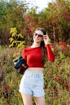 Woman holds a camera while standing in a field with colorful plants and trees