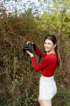 Woman holding camera in outdoor setting with plants and red berries nearby