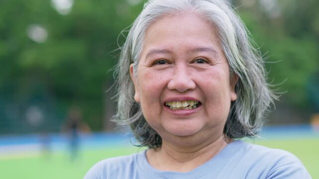 Smiling older woman faces the camera outdoors on a green sports field, framed against softly blurred trees and open space for positive wellness and active aging concepts