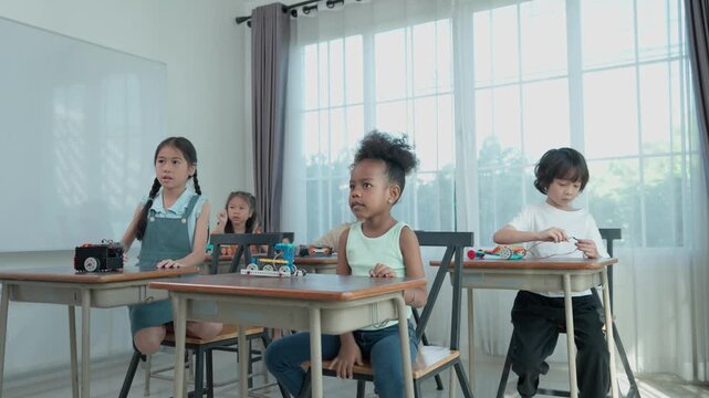 Schoolchildren sit at desks in a bright classroom, raising their hands during a group learning activity and showing active participation in education.