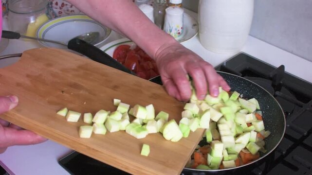 A woman prepares zucchini caviar by adding chopped zucchini to fried onions and carrots. Fresh tomatoes are also present on the table. The cooking takes place in a kitchen