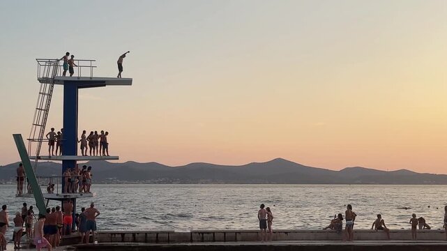 silhouette of teenager filming young man diving off a 10-meter high concrete platform into water at a beach against sunset sky located in Zadar, Croatia