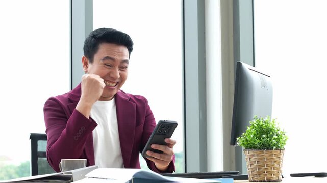 Happy Asian man uses a smartphone at an office desk and celebrates with excited gestures, smiling and raising his fists beside a computer monitor in a bright workplace.