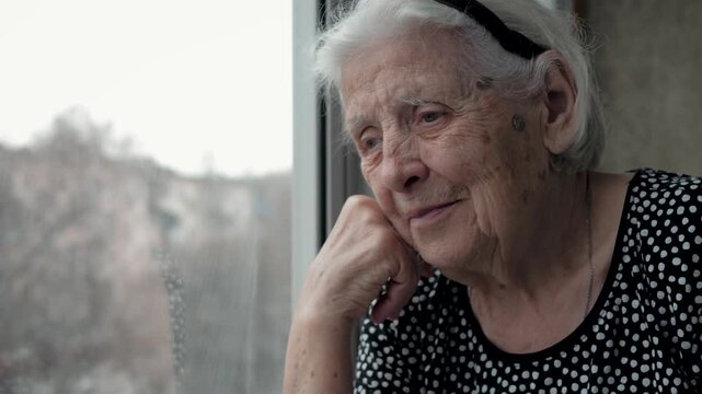 Nostalgic elderly woman with grey hair sitting alone and looking out the window. Old female remembering past with gentle smile on wrinkled face. Portrait sad grandmother feeling lonely